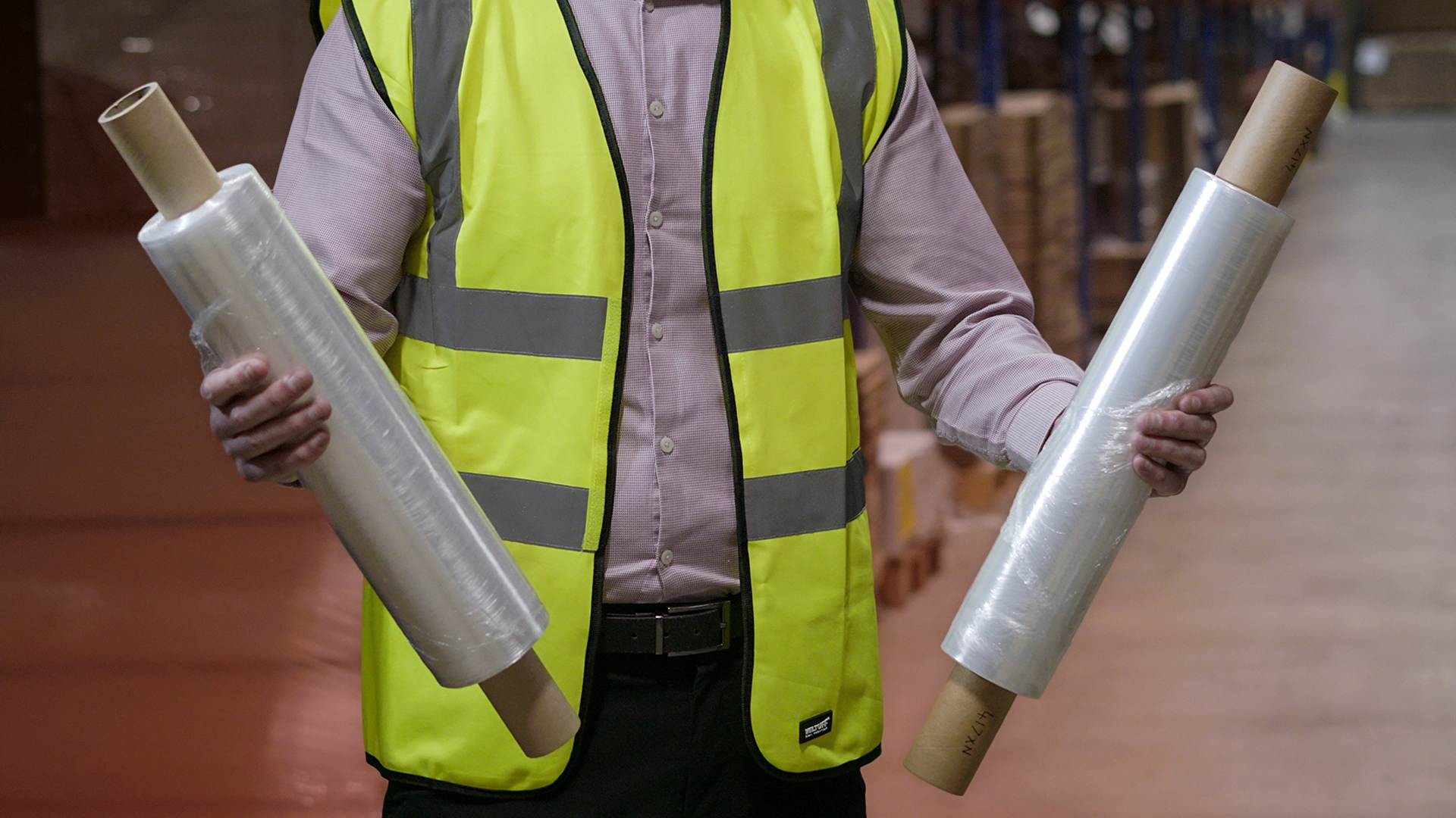 Man wearing high visibility vest holds a roll of pre stretched film in one hand and a roll of standard in the other.